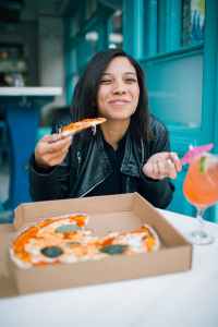 A happy young woman in a leather jacket eating a margarita pizza from a box with a cocktail that has an umbrella in it.